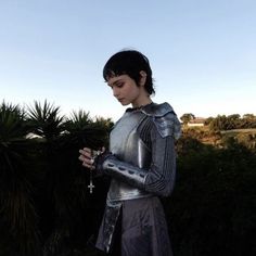 a woman dressed in armor is holding a cross and looking at her cell phone while standing outside