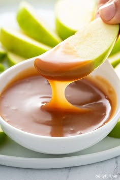 an apple being dipped with caramel sauce in a white bowl