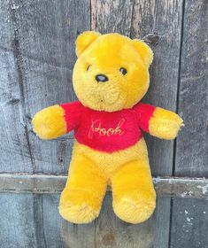 a yellow teddy bear with a red t - shirt on sitting against a wooden fence