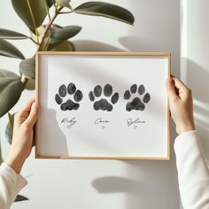 a person holding up a framed photo with two paw prints on it and a plant in the background