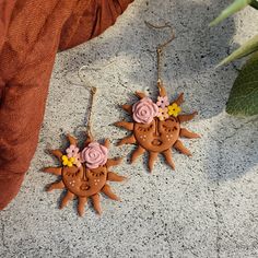 two sun shaped earrings with pink flowers on them sitting next to a leafy plant