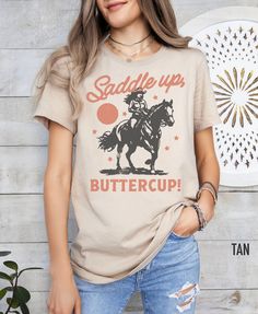 Embrace your inner cowgirl with this charming Western-inspired shirt. Featuring a playful "Saddle Up, Buttercup" slogan, this shirt is perfect for adding a touch of country flair to your wardrobe. Whether you're hitting the rodeo or just channeling some frontier spirit, this shirt is sure to make you feel like a true Western belle. We use professional quality DGT printing on all our apparel. Direct-to-garment, or DTG, is a high quality printing method that sprays ink directly onto the garment so Cute Cotton T-shirt With Cow Print, Short Sleeve Cotton Tops With Cow Print, Summer Cow Print Short Sleeve Tops, Spring Cow Print Short Sleeve Top, Cute Short Sleeve Cow Print T-shirt, Cute Cow Print Short Sleeve T-shirt, Cute Cow Print Cotton T-shirt, Rodeo Tshirt, Western Tshirt