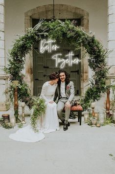 a bride and groom sitting on a bench in front of a door with greenery