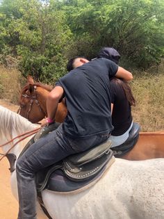 two people riding on the back of horses down a dirt road with trees in the background