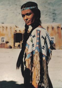 a native american woman standing in front of an adobe - style building and looking at the camera