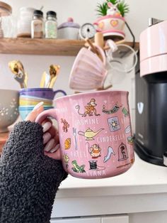 a person holding a pink coffee mug in front of a shelf with other kitchen items