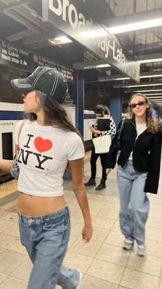 two women in jeans and baseball caps are walking through the subway station, one is wearing a t - shirt that says i love ny