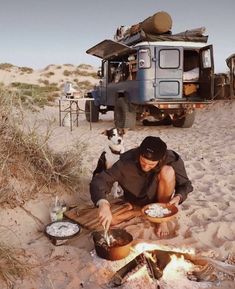 a man sitting in the sand next to a campfire with his dog looking on