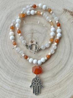 A pendant comprised of a silver plated Hamsa pendant and faceted Carnelian bead (2.5'' x 0.875'') is framed by shell, Fossil Jasper and dyed Quartz beads strung on 0.46 mm flexible wire. This necklace is 19.5'' in length with a silver plated toggle clasp. Nickel and Lead Free.  ------------------------------ All of my jewelry pieces come in a stylish drawstring pouch, ready to wear or gift! Items will ship within 1-3 business days after an order is placed and are always packed with care & love in a pet-friendly home.  **Note** Jewelry items are not waterproof, therefore it is suggested to remove prior to bathing, swimming, etc. -------------------------------- Thanks for viewing this item!  Please contact me with any questions!  I do custom orders! ~Rachel~ To see more handmade jewelry, yo White Beaded Pendant Necklace With Gemstones, Spiritual White Beaded Pendant Necklace, White Gemstone Beaded Pendant Necklace, White Gemstone Pendant Beaded Necklace, Silver Carnelian Beaded Necklace With Gemstone Beads, White Agate Beaded Necklace With Gemstone, White Gemstone Beads For Jewelry Making, White Agate Beaded Necklaces With Natural Stones, Handmade White Beads Gems And Cabochons For Gifts