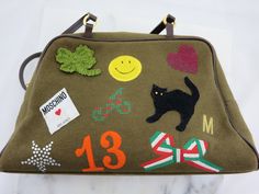 "An authentic vintage Moschino handbag, featuring \"lucky\" symbols on green felt, with brown straps. Super rare, almost impossible to find, design. Marked \"Moschino, Made in Italy\" on tag inside. There is fading and some discoloration spots to the felt. Some wear to the straps, and a slight musty smell from storage. Needs cleaning. I have priced accordingly. Measures about 12\" long, 3.5\" wide at the bottom, and about 7\" tall not including the straps. Red satin interior with one zippered si Scotland Souvenirs, Vintage Designer Handbags, Vintage Moschino, Lucky Symbols, Crazy Patchwork, Patchwork Bags, Designer Handbag, Bag Vintage, Red Satin