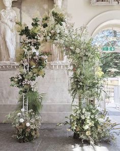 an arrangement of flowers and greenery on display in front of a wall with statues