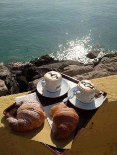 two cups of coffee and some pastries on a tray near the water's edge