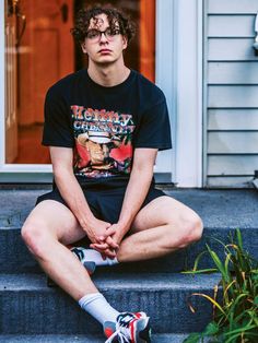 a young man sitting on steps with his legs crossed and looking at the camera while wearing glasses