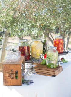 several jars filled with different types of drinks on a white table cloth under a tree