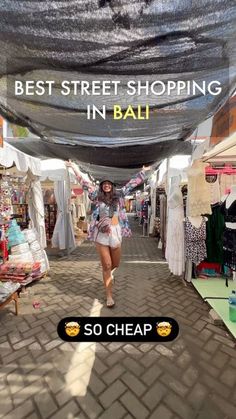 a woman walking down a street next to tents with clothes hanging from the ceiling and text reading best street shopping in bali so cheap