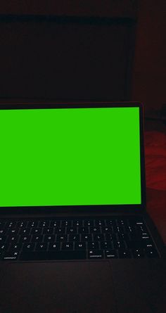 an open laptop computer with a green screen on a dark surface in front of a bed