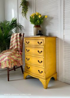 a yellow dresser sitting next to a chair and potted plant on top of it