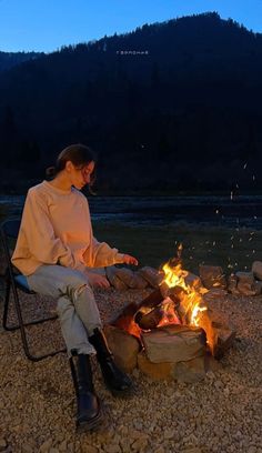 a woman sitting in a chair next to a campfire at night with mountains in the background