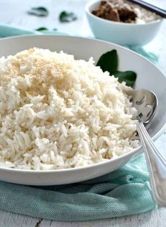 a white bowl filled with rice on top of a blue and white table cloth next to silver spoons