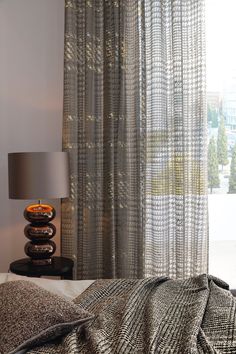 a bed sitting next to a window covered in gray and white curtains, with a lamp on top of it