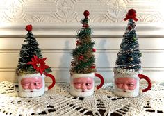 three ceramic mugs with christmas trees and santa's faces on them sitting on a doily