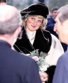 a woman wearing a black hat and fur coat holding a flower in her right hand