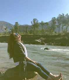 a woman sitting on top of a rock next to a river