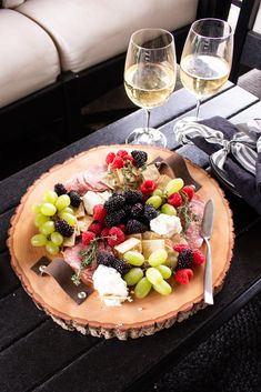 a wooden plate topped with fruit and cheese next to two wine glasses