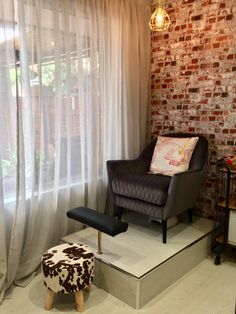 a chair and foot stool in front of a brick wall with white drapes on it