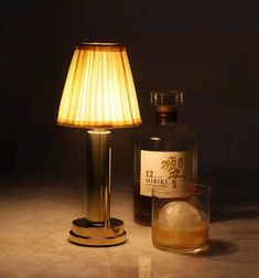 a bottle and glass sitting on a table next to a lamp that is turned on