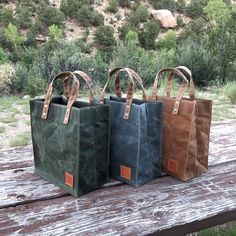 Three small waxed canvas totes on a picnic table in a park. Handcrafted byRoad Dirt and Pie and available in green, gray, and brown colors. Everyday Bag With Reinforced Handles In Waxed Canvas, Waxed Canvas Tote Bags With Reinforced Handles, Practical Rectangular Canvas Bag With Waxed Finish, Practical Waxed Canvas Bag, Practical Rectangular Waxed Canvas Bag, Eco-friendly Everyday Waxed Canvas Bag, Rectangular Waxed Canvas Bag With Top Carry Handle, Everyday Waxed Canvas Bag With Reinforced Handles, Practical Rectangular Bag With Waxed Finish