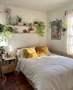 a bed with white sheets and yellow pillows in a bedroom next to two potted plants