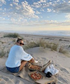 a woman sitting on top of a sandy beach next to a basket filled with pizza