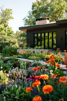 a garden with lots of different flowers and plants in front of a house that is surrounded by trees