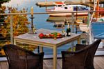 there are two chairs and a table on the deck by the water with boats in the background