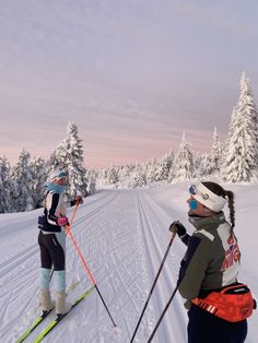 two people on skis in the snow with trees behind them and pink sky above