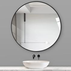 a round mirror hanging on the wall above a white sink and black faucet