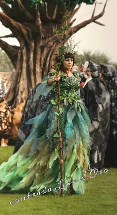 a woman dressed in green and blue standing next to a tree