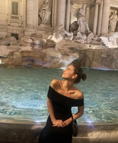 a woman sitting on the edge of a fountain in front of a building with statues