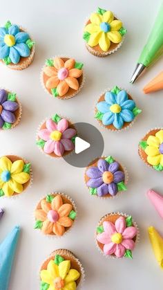 cupcakes decorated with icing and flowers are arranged on a white surface next to colored markers