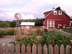 a red barn with a windmill and flowers in the foreground, surrounded by a fence