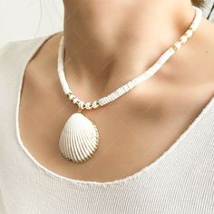 Experience the charm of the ocean with our Natural Scallop Shell Necklace. This meticulously handcrafted piece features a large, authentic scallop shell pendant, elegantly trimmed in gold, providing a striking centerpiece. The necklace is strung with round, white ceramic beads, interspersed with golden accents that enhance its sophistication. Perfect for those who love to incorporate elements of nature into their style, this necklace promises versatility and elegance, making it an ideal gift for Handmade Pearl White Shell Necklace For Beach, Pearl White Shell Jewelry For Beach, Pearl White Beaded Jewelry For Beach, Pearl White Beaded Jewelry For The Beach, Pearl White Shell Necklace For Beach, White Gold Beaded Necklace For Beach, Gold Shell Beaded Necklaces For Beach, Pearl White Shell Jewelry For The Beach, Gold Shell Beaded Necklace For Beach