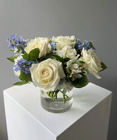 a glass vase filled with white and blue flowers on top of a table next to a gray wall