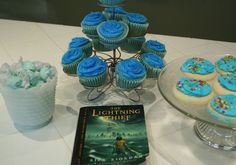 a table topped with blue frosted cupcakes next to a cake and book