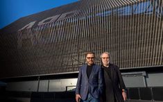 two men standing next to each other in front of a building with metal slats on it