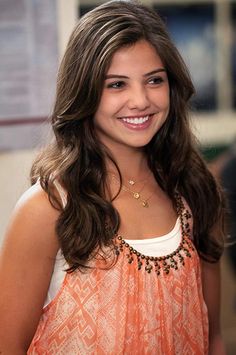 a woman with long brown hair smiling at the camera and wearing an orange top, standing in front of a bulletin board