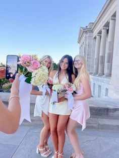 three beautiful young women standing next to each other in front of a building holding flowers