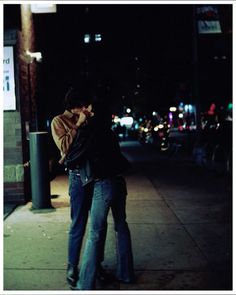 a man and woman standing next to each other on a sidewalk at night with lights in the background