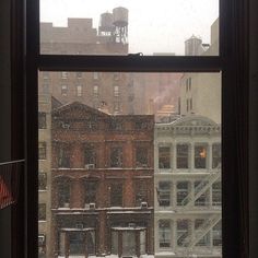 a window with snow falling on the ground and buildings in the backgrouds