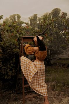 a woman in a yellow and white checkered dress is standing on a ladder near an apple tree